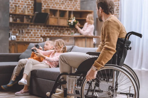 Padre en silla de ruedas mirando lo felices que son los niños jugando videojuegos - foto de stock