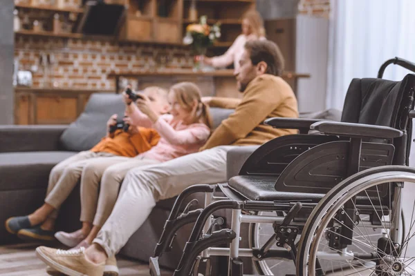 Padre mirando cómo los niños juegan videojuegos con silla de ruedas en primer plano - foto de stock