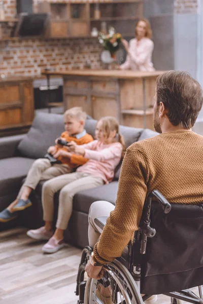 Pai na cadeira de rodas olhando como as crianças jogando videogame — Fotografia de Stock