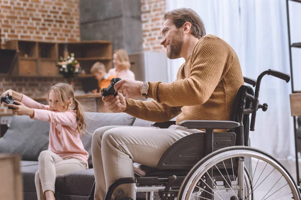 Happy daughter and father on wheelchair playing video game with joysticks — Stock Photo