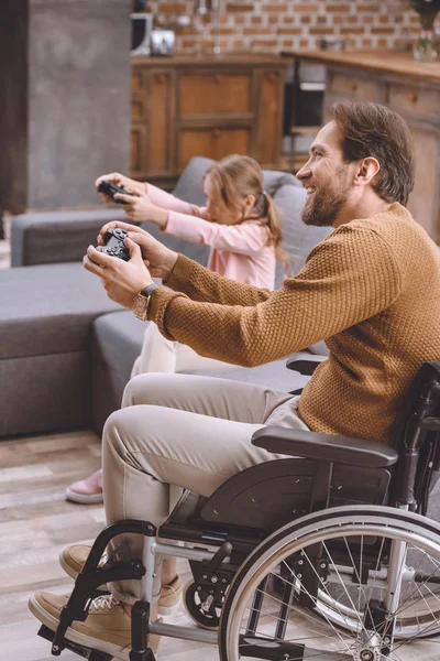 Feliz hija y padre en silla de ruedas jugando videojuego con joysticks - foto de stock