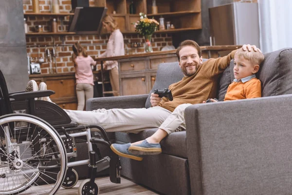 Heureux père handicapé avec les jambes sur fauteuil roulant et mignon petit fils jouer avec joysticks ensemble à la maison — Photo de stock