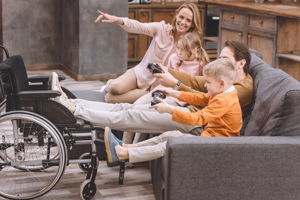 Família feliz com pai deficiente colocando pernas em cadeira de rodas e brincando com joysticks em casa — Fotografia de Stock
