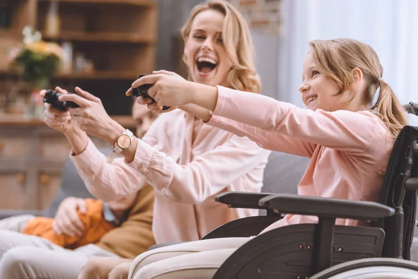 Família alegre com criança com deficiência em cadeira de rodas brincando com joysticks juntos em casa — Fotografia de Stock