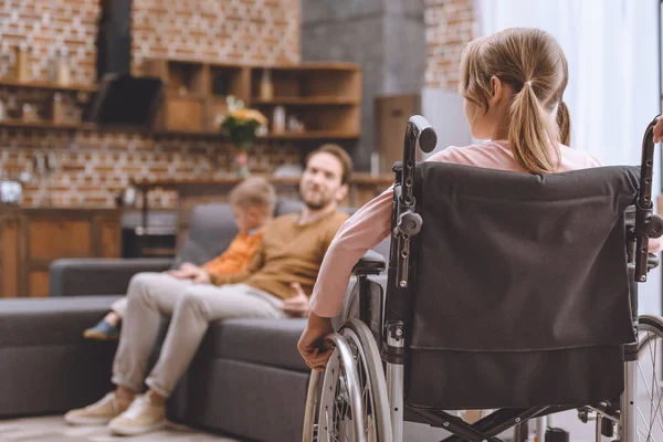 Vista trasera del niño discapacitado sentado en silla de ruedas y mirando al padre y al hermano sentados en el sofá en casa - foto de stock