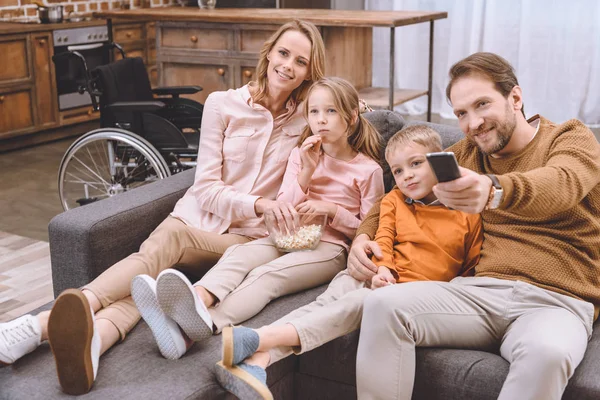 Щаслива сім'я сидить на дивані і дивиться телевізор разом та інвалідне крісло, стоячи позаду — стокове фото