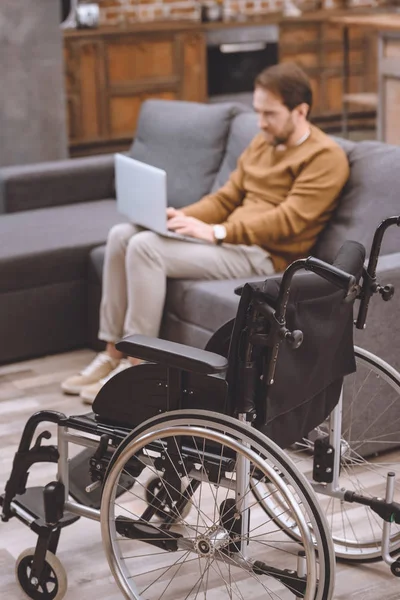 Vista ravvicinata della sedia a rotelle e dell'uomo disabile che utilizza il computer portatile sul divano a casa — Foto stock