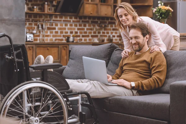 Mujer feliz mirando marido discapacitado poner las piernas en silla de ruedas y el uso de la computadora portátil en casa — Stock Photo