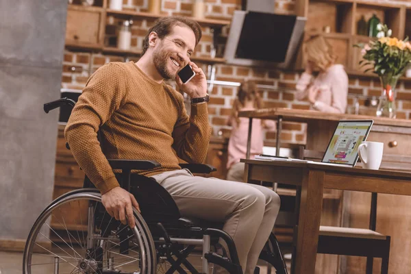 Homem deficiente feliz na cadeira de rodas falando no smartphone e usando laptop em casa — Fotografia de Stock