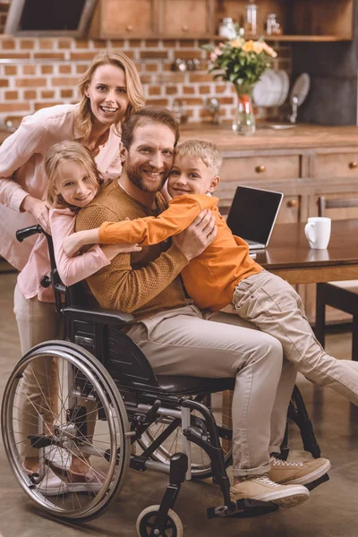 Glückliche Familie mit zwei Kindern und Vater im Rollstuhl, die sich zu Hause umarmen und in die Kamera lächeln — Stockfoto