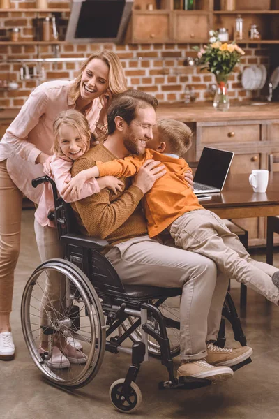 Glückliche Familie mit zwei Kindern und Vater im Rollstuhl, die sich zu Hause umarmen — Stockfoto