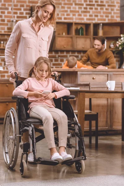 Sonriente madre mirando a la hija discapacitada sentada en silla de ruedas y usando un teléfono inteligente en casa - foto de stock