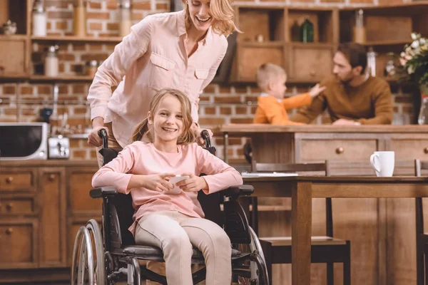 Mother looking at smiling disabled daughter sitting in wheelchair and using smartphone at home — Stock Photo