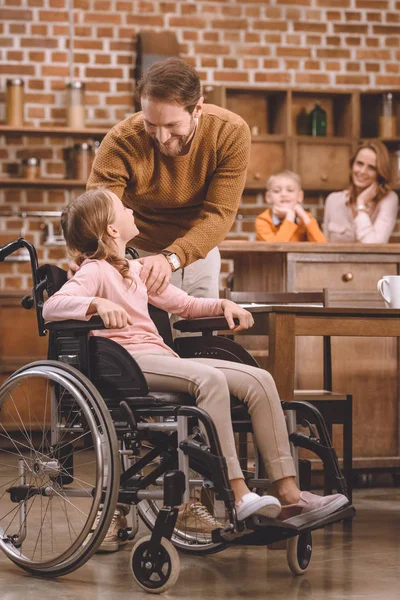 Pai e linda filhinha em cadeira de rodas sorrindo uns aos outros em casa — Fotografia de Stock