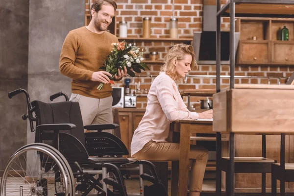 Side view of disabled woman taking notes and smiling man holding bouquet of flowers at home — Stock Photo