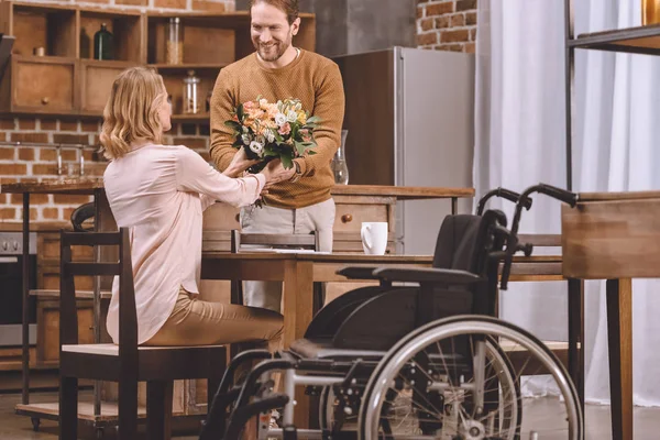 Homem sorrindo apresentando flores para a esposa deficiente e cadeira de rodas de pé por perto — Fotografia de Stock