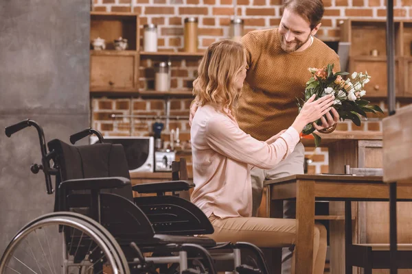Улыбающийся мужчина дарит цветы жене-инвалиду дома — стоковое фото