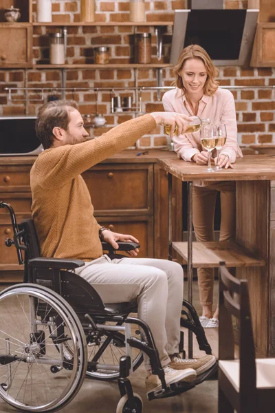 Mann im Rollstuhl schenkt Wein ein, während lächelnde Frau Gläser zu Hause hält — Stockfoto