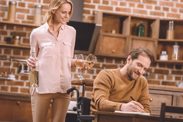 Donna sorridente che tiene bicchieri di vino e bottiglia mentre l'uomo in sedia a rotelle prende appunti a casa — Foto stock