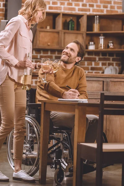Schnappschuss einer lächelnden Frau mit Weingläsern und Flasche, während ihr Mann im Rollstuhl zu Hause Notizen macht — Stockfoto