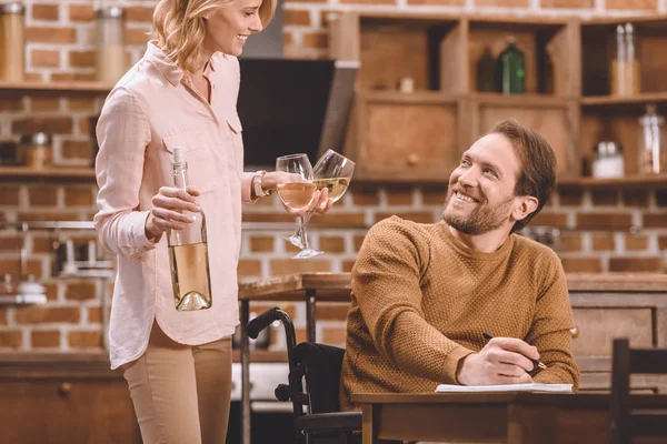 Улыбающаяся женщина держит бокалы и бутылку вина, а муж инвалид в инвалидной коляске делает заметки дома — стоковое фото