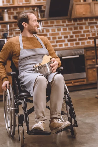 Homme handicapé souriant dans une casserole en fauteuil roulant avec des spaghettis crus et regardant loin à la maison — Photo de stock