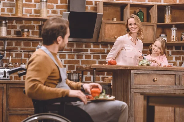 Feliz madre e hija mirando a padre sentado en silla de ruedas y cortando verduras para la cena - foto de stock