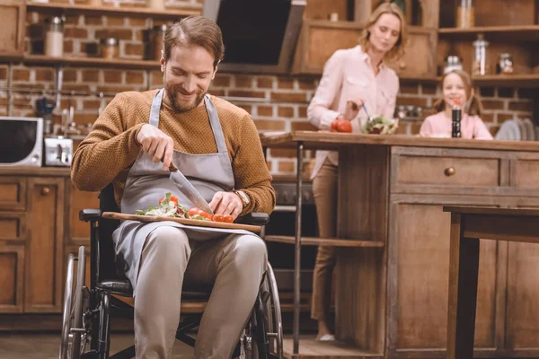 Uomo sorridente in sedia a rotelle tagliare verdure per insalata mentre felice madre e figlia in piedi dietro — Foto stock