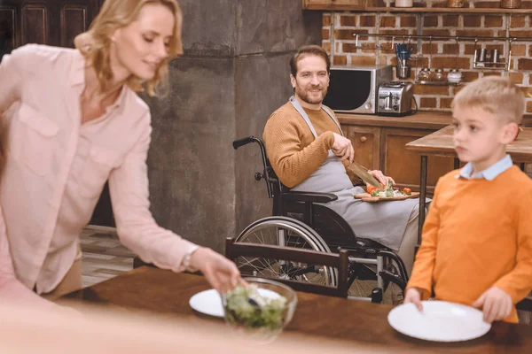 Madre con hijo sirviendo mesa para la cena mientras padre en silla de ruedas cortando verduras en casa - foto de stock