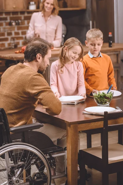 Crianças felizes com o pai na mesa de serviço de cadeira de rodas para o jantar em casa — Fotografia de Stock
