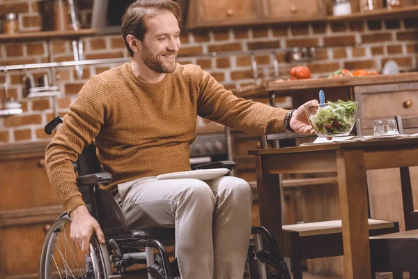 Homem com deficiência sorridente em cadeira de rodas segurando tigela de vidro de salada de legumes em casa — Fotografia de Stock