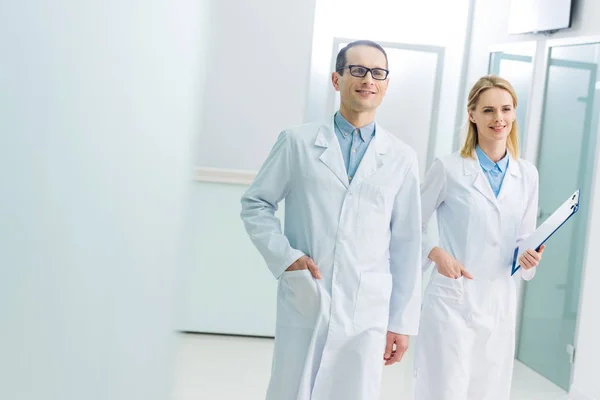 Médicos sonrientes con batas blancas con diagnóstico en el pasillo del hospital - foto de stock