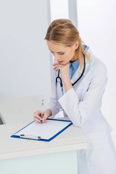 Mujer rubia médico escribiendo diagnóstico en el hospital - foto de stock