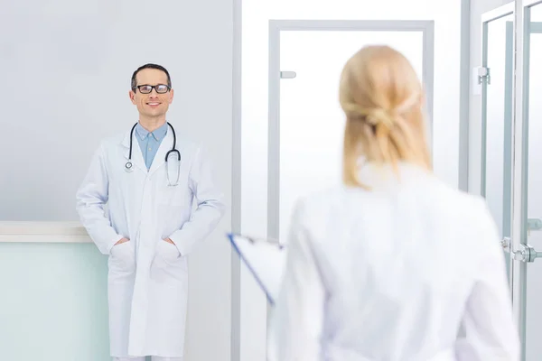 Médico varón de bata blanca con estetoscopio mirando a su colega en la clínica - foto de stock