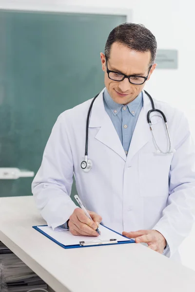 Улыбающийся доктор в белом халате со стетоскопом пишет диагноз в больнице — стоковое фото
