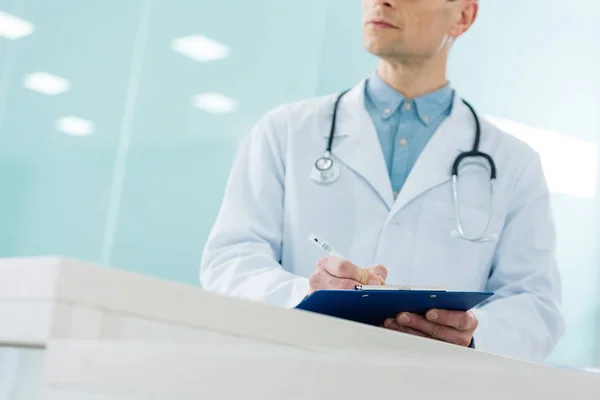 Cropped view of male doctor in white coat with stethoscope writing diagnosis in hospital — Stock Photo