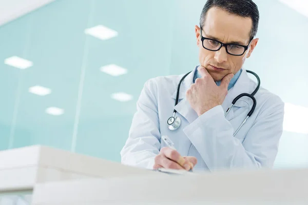 Вдумчивый доктор в белом халате со стетоскопом пишет диагноз в больнице — стоковое фото