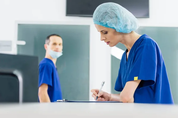 Cirujano femenino escribiendo diagnóstico mientras su colega la mira en el hospital - foto de stock