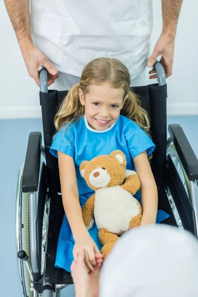Улыбающийся ребенок сидит с мягкой игрушкой на инвалидной коляске и смотрит в камеру — стоковое фото