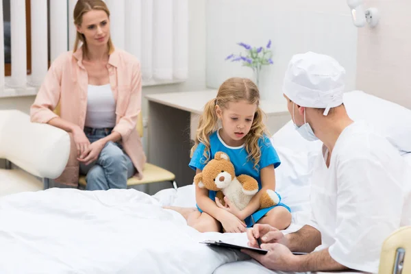 Médico hablando con el niño y sentado en la cama del hospital - foto de stock
