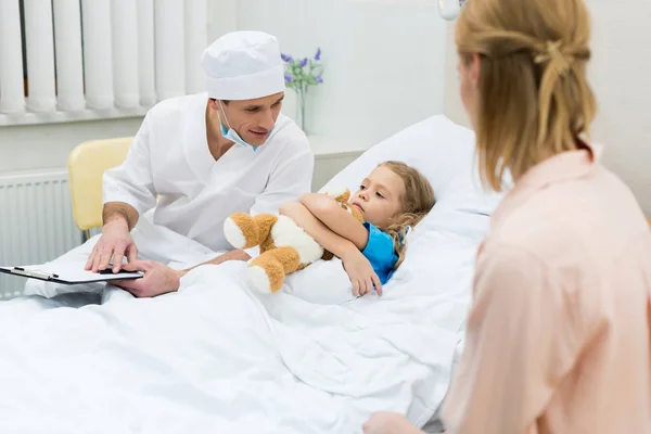 Médecin assis avec presse-papiers et parler avec enfant patient — Photo de stock