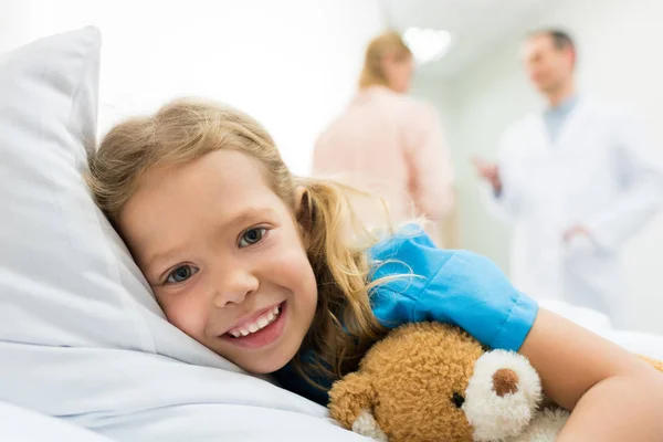 Criança feliz deitado na cama do hospital, enquanto a mãe falando com o médico — Fotografia de Stock