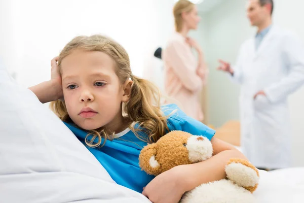 Niño molesto acostado en la cama del hospital mientras la madre habla con el médico - foto de stock