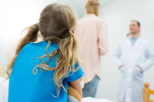 Rear view of kid sitting on hospital bed while mother talking with doctor — Stock Photo