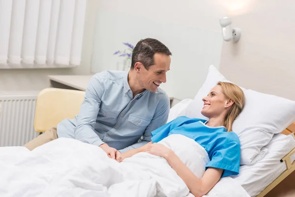 Sonriente marido sentado en esposa cama en hospital - foto de stock