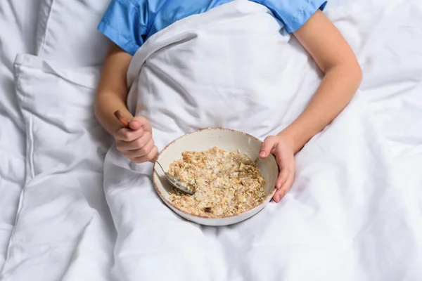 Image recadrée d'un enfant d'âge préscolaire couché sur son lit à l'hôpital avec assiette de flocons d'avoine — Photo de stock