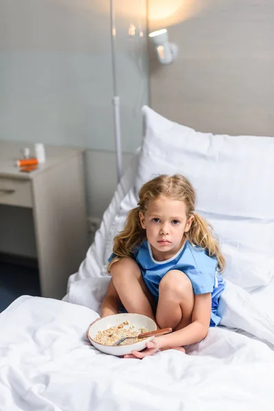 Criança pré-escolar sentado na cama no hospital com prato de aveia — Fotografia de Stock