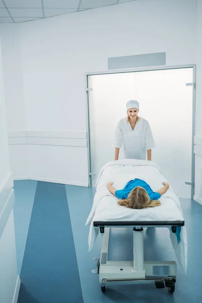 Smiling doctor moving preschooler patient on medical stretcher in corridor — Stock Photo