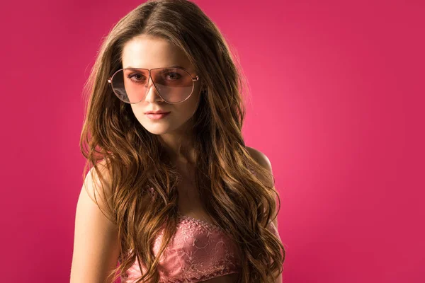 Портрет привабливої дівчини в сонцезахисних окулярах і бюстгальтер дивиться на камеру ізольовано на рожевому — стокове фото