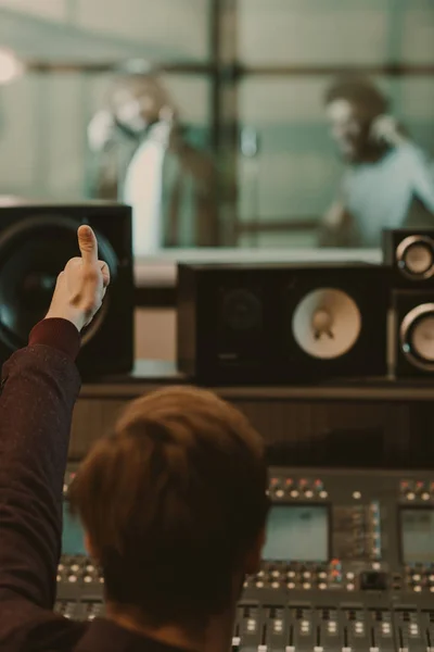 Productor de sonido mostrando el pulgar hacia arriba a los cantantes detrás de vidrio en el estudio de grabación - foto de stock
